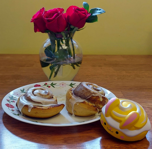 On a wooden table sits a vase of roses, a plate with two cinnamon buns, and a cinnamon bunny slow rise squishy.