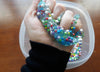 A person's hand squeezes blue slime with foam beads. Below, on a wood surface is a container filled with excess foam beads.