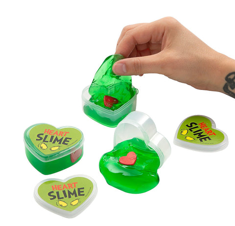 Three containers of green Grinchmas slime. On the lids is the text "Heart Slime" and the eyes of the Grinch character. Inside each slime is a red styrofoam heart. One container is closed. One container is open, and slime spills out. The last container is open and a person's hand stretches slime out of it.