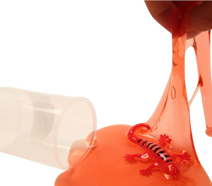 Container of slime open with red slime pouring out onto a surface. A red lizard sits in the slime. A person's hand stretches the slime upward.